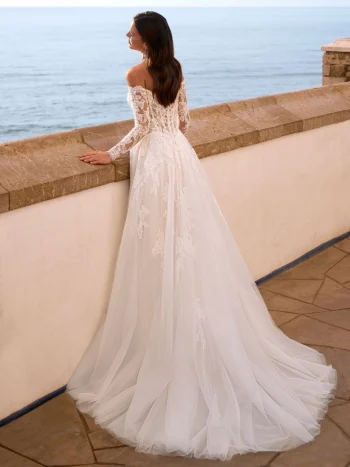 A woman in the FAIZA by Love by Enz white, off-the-shoulder lace wedding dress stands by a stone wall overlooking the ocean—a vision of elegance and romance.