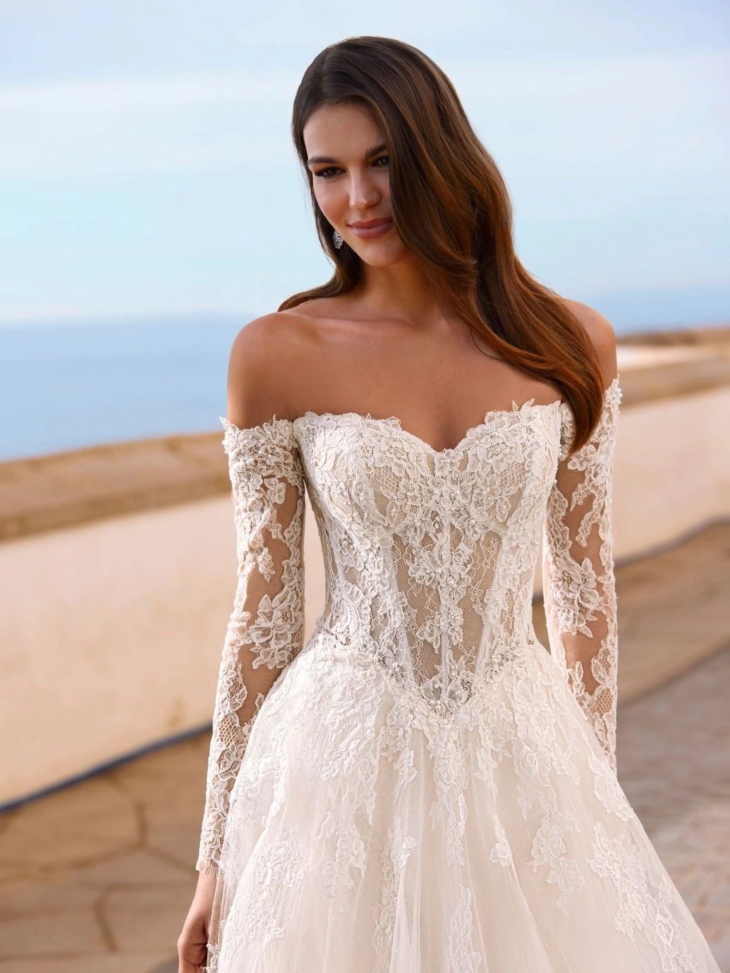 A woman wearing the FAIZA by Love by Enz off-the-shoulder, long-sleeve lace wedding dress stands outdoors with a blurred ocean and stone railing in the background. A woman wearing the FAIZA by Love by Enz off-the-shoulder, long-sleeve lace wedding dress stands outdoors with a blurred ocean and stone railing in the background.