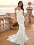 A woman in an off-shoulder, fitted white FIE by Love by Enz gown with a train stands on a terrace overlooking the ocean at sunset.