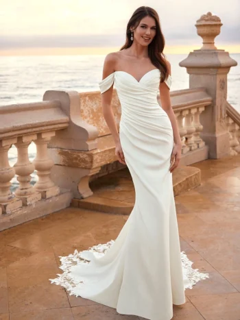A woman in an off-shoulder, fitted white FIE by Love by Enz gown with a train stands on a terrace overlooking the ocean at sunset.