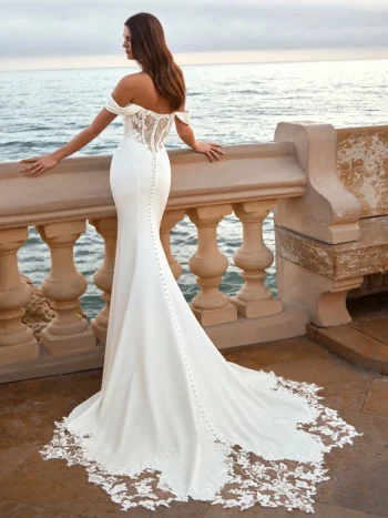 Woman in the FIE by Love by Enz off-shoulder, fitted white bridal gown with a lace train stands by a stone balustrade overlooking the ocean at sunset.