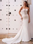 A woman in the FOSIRA by Love by Enz strapless fitted wedding dress, featuring a lace bodice and long train, stands against a white door with black decorative accents.