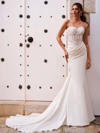 A woman in the FOSIRA by Love by Enz strapless fitted wedding dress, featuring a lace bodice and long train, stands against a white door with black decorative accents.