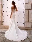 A woman in the FOSIRA by Love by Enz strapless fitted wedding dress, featuring a lace bodice and long train, stands against a white door with black decorative accents.