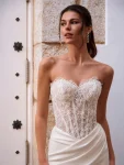 A woman in the FOSIRA by Love by Enz strapless fitted wedding dress, featuring a lace bodice and long train, stands against a white door with black decorative accents.