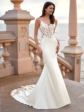 Woman wearing a fitted white wedding dress with lace bodice and train, standing on a stone terrace by the sea.