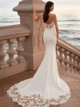 Woman wearing a fitted white wedding dress with lace bodice and train, standing on a stone terrace by the sea.
