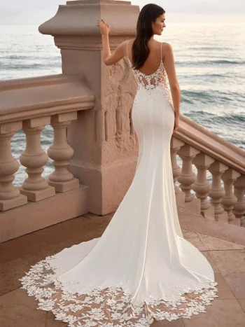 A woman in a white, sleeveless, lace-trimmed wedding dress stands on stone steps overlooking the ocean at sunset.