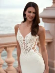 Woman wearing a fitted white wedding dress with lace bodice and train, standing on a stone terrace by the sea.