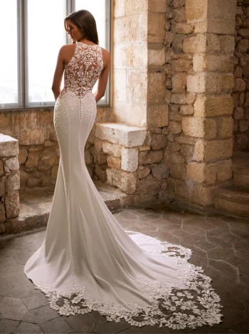 A woman in a white lace GRACY by Etoile Enz mermaid wedding dress with a long train stands by a stone wall and window, facing away from the camera.