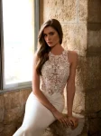 A woman wearing the GRACY by Etoile Enz wedding gown—a fitted, sleeveless white dress adorned with lace details—stands near a stone wall and window, radiating timeless elegance.