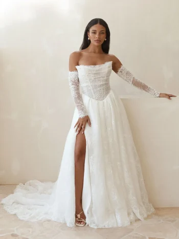 Serene and graceful, the woman stands against a neutral wall in the LILAC/SR2641 by Serene gown—an off-the-shoulder white lace wedding dress featuring sheer long sleeves, a thigh-high slit, and a flowing floor-length train. Subtle lilac undertones enhance the romantic elegance of this captivating bridal look.