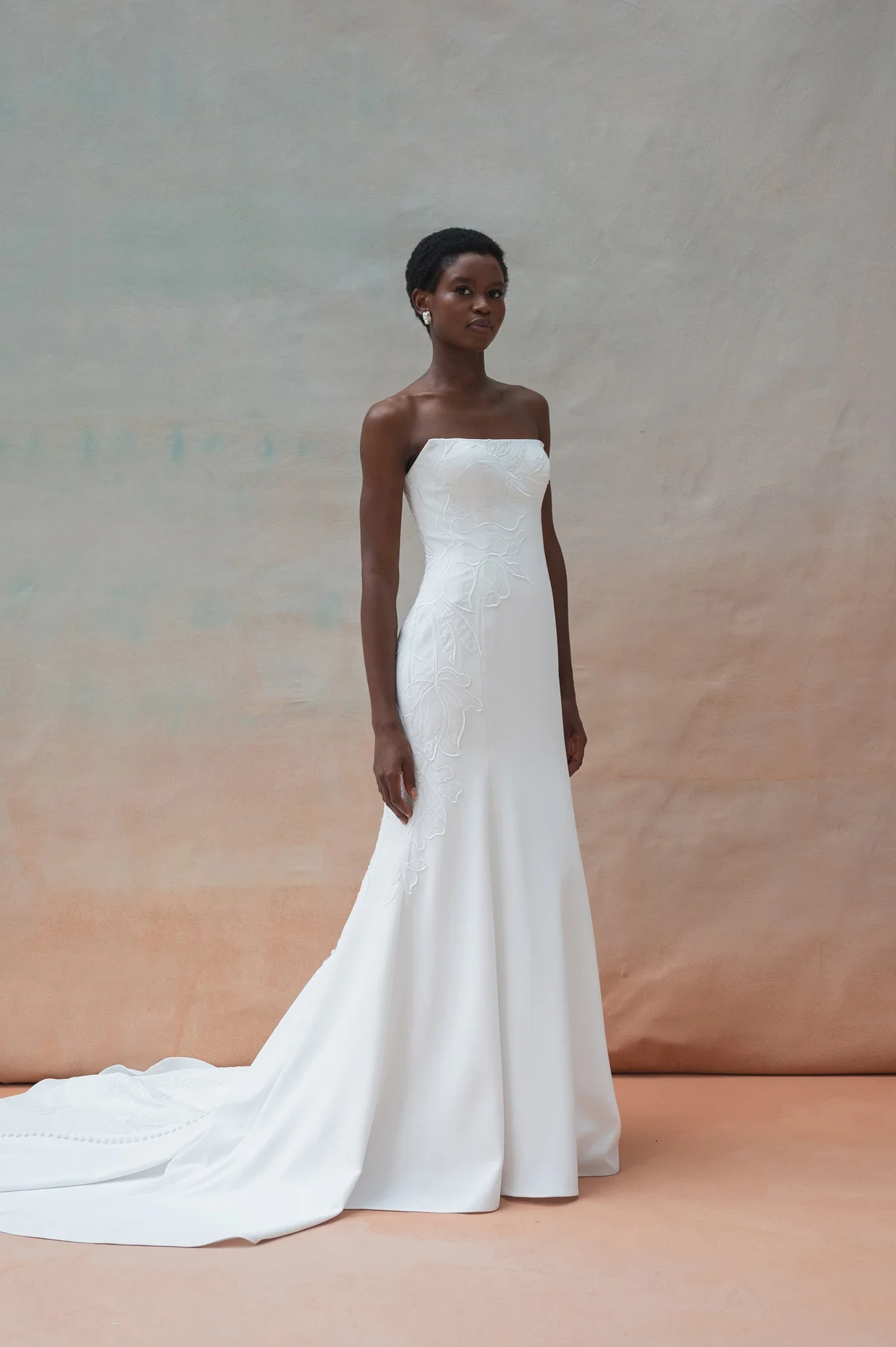 A bridal elegance is captured as a woman wearing the TRICIA by Jenny Yoo strapless white wedding gown with a long train stands against a neutral, softly colored backdrop. A bridal elegance is captured as a woman wearing the TRICIA by Jenny Yoo strapless white wedding gown with a long train stands against a neutral, softly colored backdrop.