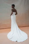 A bridal elegance is captured as a woman wearing the TRICIA by Jenny Yoo strapless white wedding gown with a long train stands against a neutral, softly colored backdrop.