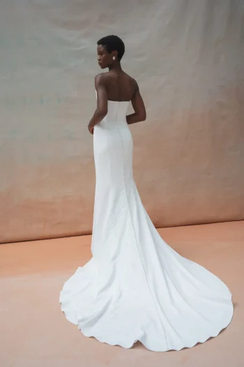 A woman stands facing away against a neutral backdrop, wearing the TRICIA by Jenny Yoo, a strapless white bridal gown with a long train.