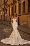 A woman wearing the B1438 by Joelle Olivia, a strapless fitted white bridal gown, stands on a cobblestone street in front of historic yellow buildings.