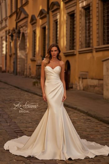 A woman wearing the B1438 by Joelle Olivia, a strapless fitted white bridal gown, stands on a cobblestone street in front of historic yellow buildings.