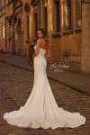 A woman wearing the B1438 by Joelle Olivia, a strapless fitted white bridal gown, stands on a cobblestone street in front of historic yellow buildings.