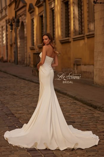 A woman wearing the B1438 by Joelle Olivia, a strapless fitted white bridal gown with a long train and button details down the back, stands on a cobblestone street and looks over her shoulder.