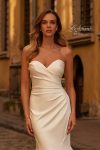 A woman wearing the B1438 by Joelle Olivia, a strapless fitted white bridal gown, stands on a cobblestone street in front of historic yellow buildings.