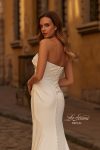 A woman wearing the B1438 by Joelle Olivia, a strapless fitted white bridal gown, stands on a cobblestone street in front of historic yellow buildings.