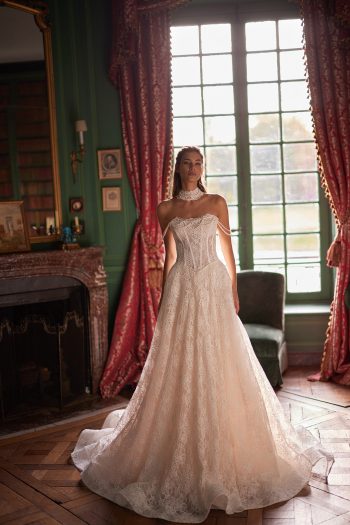 Woman in a strapless lace wedding gown stands in an elegant room with large windows, red curtains, and a fireplace.