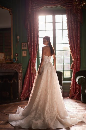 A woman wearing the CORDELIA strapless lace wedding gown stands in front of a tall window with red curtains in a vintage-style room.