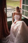 Woman in a strapless lace wedding gown stands in an elegant room with large windows, red curtains, and a fireplace.