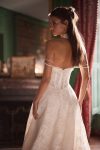 Woman in a strapless lace wedding gown stands in an elegant room with large windows, red curtains, and a fireplace.