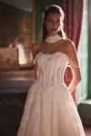 Woman in a strapless lace wedding gown stands in an elegant room with large windows, red curtains, and a fireplace.