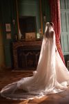 Woman in a strapless lace wedding gown stands in an elegant room with large windows, red curtains, and a fireplace.