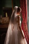 Woman in a strapless lace wedding gown stands in an elegant room with large windows, red curtains, and a fireplace.