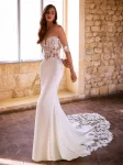 A woman wearing the GIVELLE by Etoile Enz, a fitted off-the-shoulder white wedding dress featuring intricate lace details and a long lace train, stands indoors near a stone wall and window.