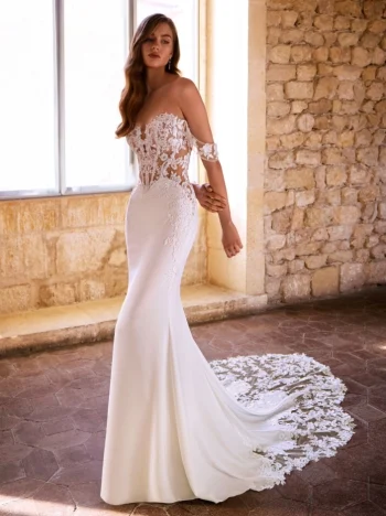 A woman wearing the GIVELLE by Etoile Enz, a fitted off-the-shoulder white wedding dress featuring intricate lace details and a long lace train, stands indoors near a stone wall and window.