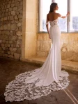 A woman wearing the GIVELLE by Etoile Enz, a fitted off-the-shoulder white wedding dress featuring intricate lace details and a long lace train, stands indoors near a stone wall and window.