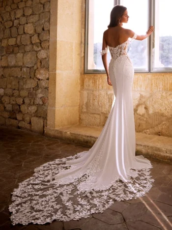 A woman wearing the GIVELLE by Etoile Enz, an off-shoulder wedding dress with a lace train, stands by a large window looking outside. The gown showcases delicate floral detailing and a fitted silhouette.