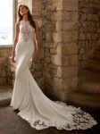 A woman wearing the GRACY by Etoile Enz wedding gown—a fitted, sleeveless white dress adorned with lace details—stands near a stone wall and window, radiating timeless elegance.