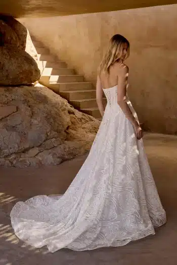 A woman in a strapless, lace wedding dress with a long train stands indoors near stone steps and a large rock.