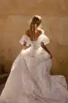 Woman in a strapless white lace wedding gown stands indoors near stone stairs and a textured wall.
