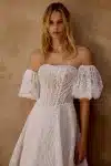 Woman in a strapless white lace wedding gown stands indoors near stone stairs and a textured wall.
