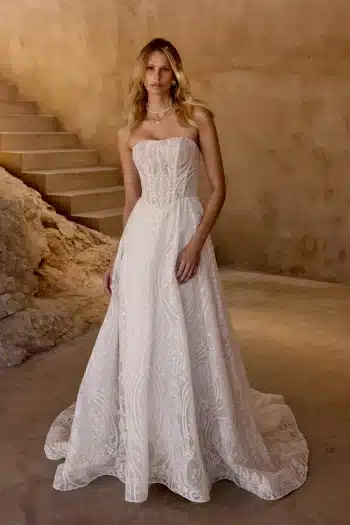 Woman in a strapless white lace wedding gown stands indoors near stone stairs and a textured wall.