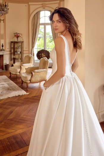 A woman in the 88504/MCKENNA by Justin Alexander, a white sleeveless gown featuring button details, stands in an elegant room with wooden floors and classic furniture.
