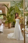 A woman wearing the 88511/MELODY by Justin Alexander, a strapless white wedding gown, stands indoors beside large potted plants. Natural light and classic decor provide an elegant and timeless backdrop for this exquisite dress.