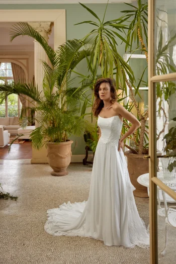 A woman wearing the 88511/MELODY by Justin Alexander, a strapless white wedding gown, stands indoors beside large potted plants. Natural light and classic decor provide an elegant and timeless backdrop for this exquisite dress.