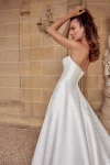 A woman in a strapless white satin 88512/MIA by Justin Alexander wedding gown stands in front of a stone statue and arched alcove, flanked by two potted plants. This exquisite bridal look radiates timeless elegance.