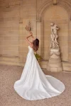 A woman in a strapless white satin 88512/MIA by Justin Alexander wedding gown stands in front of a stone statue and arched alcove, flanked by two potted plants. This exquisite bridal look radiates timeless elegance.