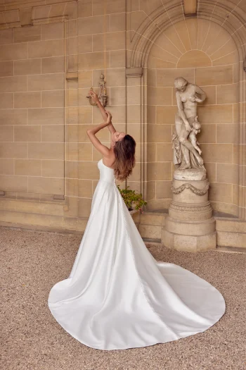 A woman in a strapless white 88512/MIA by Justin Alexander wedding dress stands outdoors, facing a stone wall and statue, holding her arms up as the gown’s long train flows behind her. The elegant scene captures the timeless beauty of this exquisite dress.