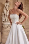 A woman in a strapless white satin 88512/MIA by Justin Alexander wedding gown stands in front of a stone statue and arched alcove, flanked by two potted plants. This exquisite bridal look radiates timeless elegance.