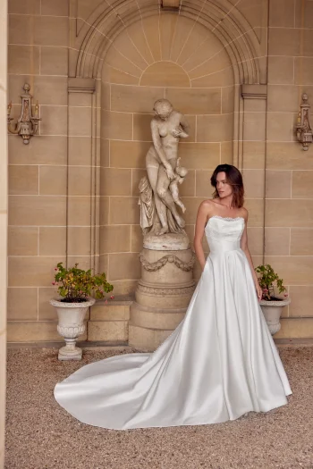 A woman in a strapless white satin 88512/MIA by Justin Alexander wedding gown stands in front of a stone statue and arched alcove, flanked by two potted plants. This exquisite bridal look radiates timeless elegance.