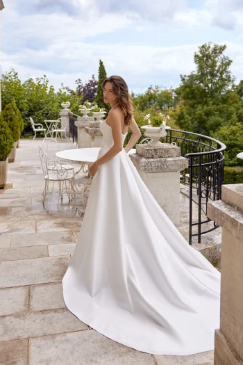 A woman in a strapless white 88515/MADISON wedding dress stands on a stone terrace, glancing over her shoulder toward a round table and chairs, with greenery in the background.